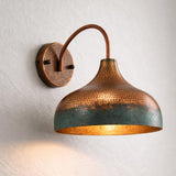 A hammered copper wall sconce with a dome-shaped shade and a warm glowing light inside, mounted on a textured off-white wall.
