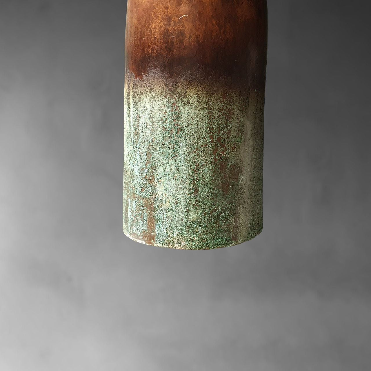 A close-up of a cylindrical ceramic vase or container with a rustic glaze, showing a dark brown top section and a textured green and brown lower section, set against a soft gray gradient background.