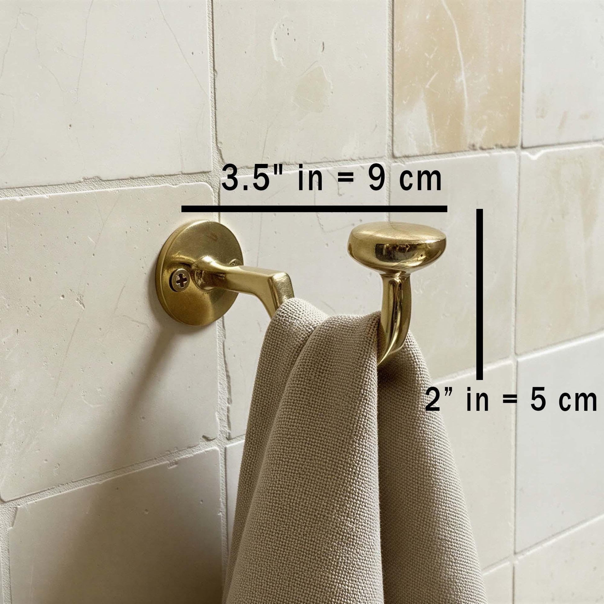 Gold towel hook on a tiled wall with measurements indicated