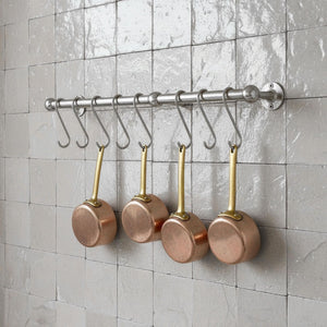 Set of copper pots with brass handles hanging on a metal rack against a tiled wall.