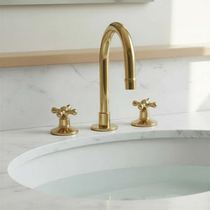 Gold faucet with two handles on a white sink against a light marble wall.