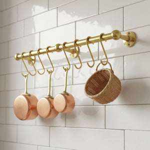 Gold hooks with copper pots and a woven basket on a white tiled wall.