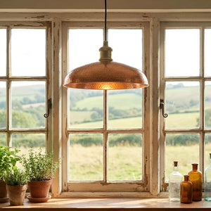 Copper pendant light hanging above a wooden window sill with plants and bottles, overlooking a scenic landscape.