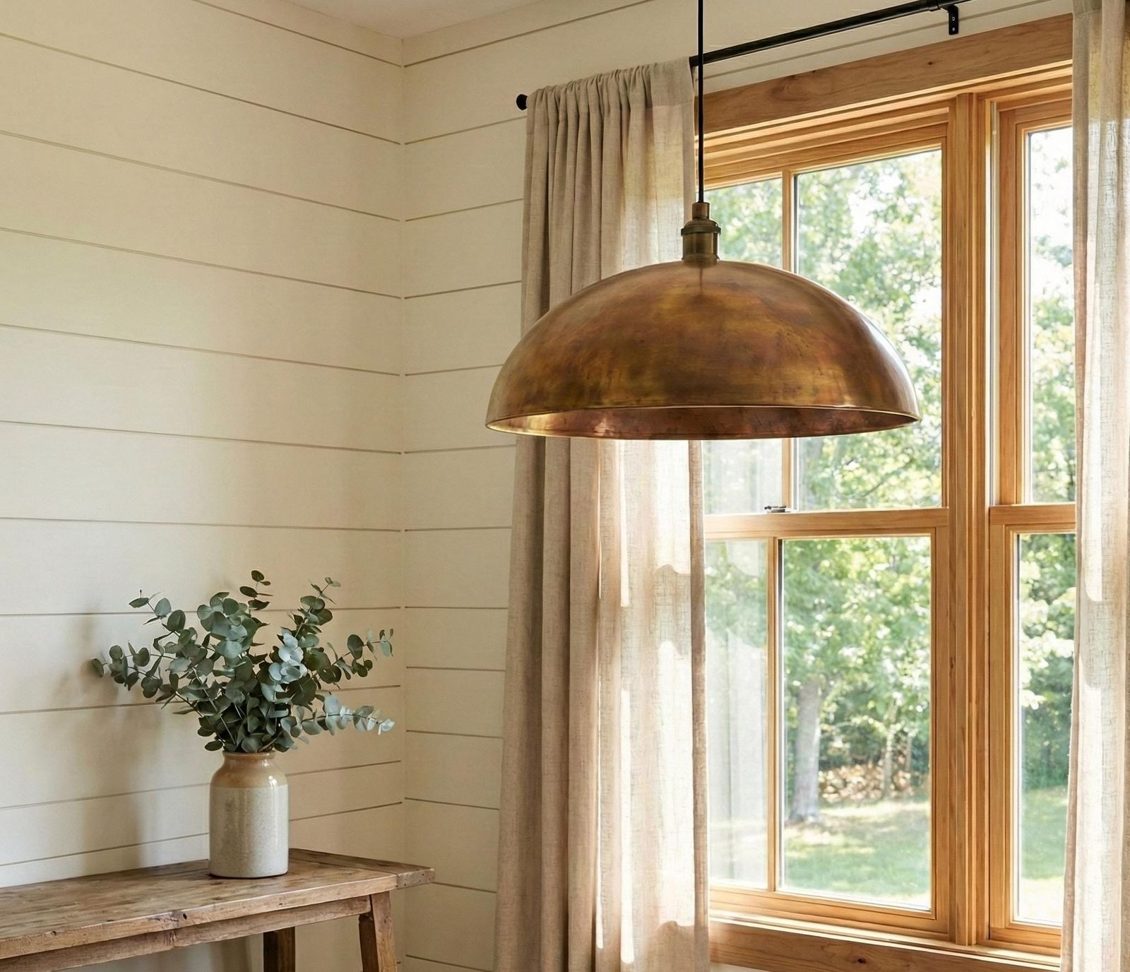 Brass pendant light hanging above a wooden table with a plant in a room with a large window.
