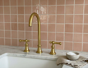 Gold faucet set on a marble countertop with pink tiled wall in the background
