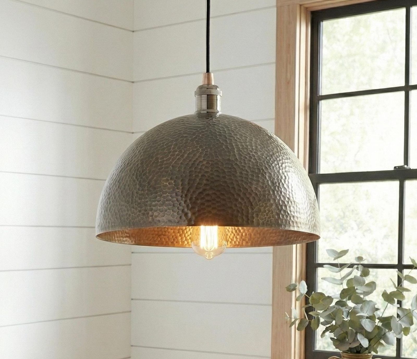 Hammered metal pendant light hanging in a room with a window and plant.