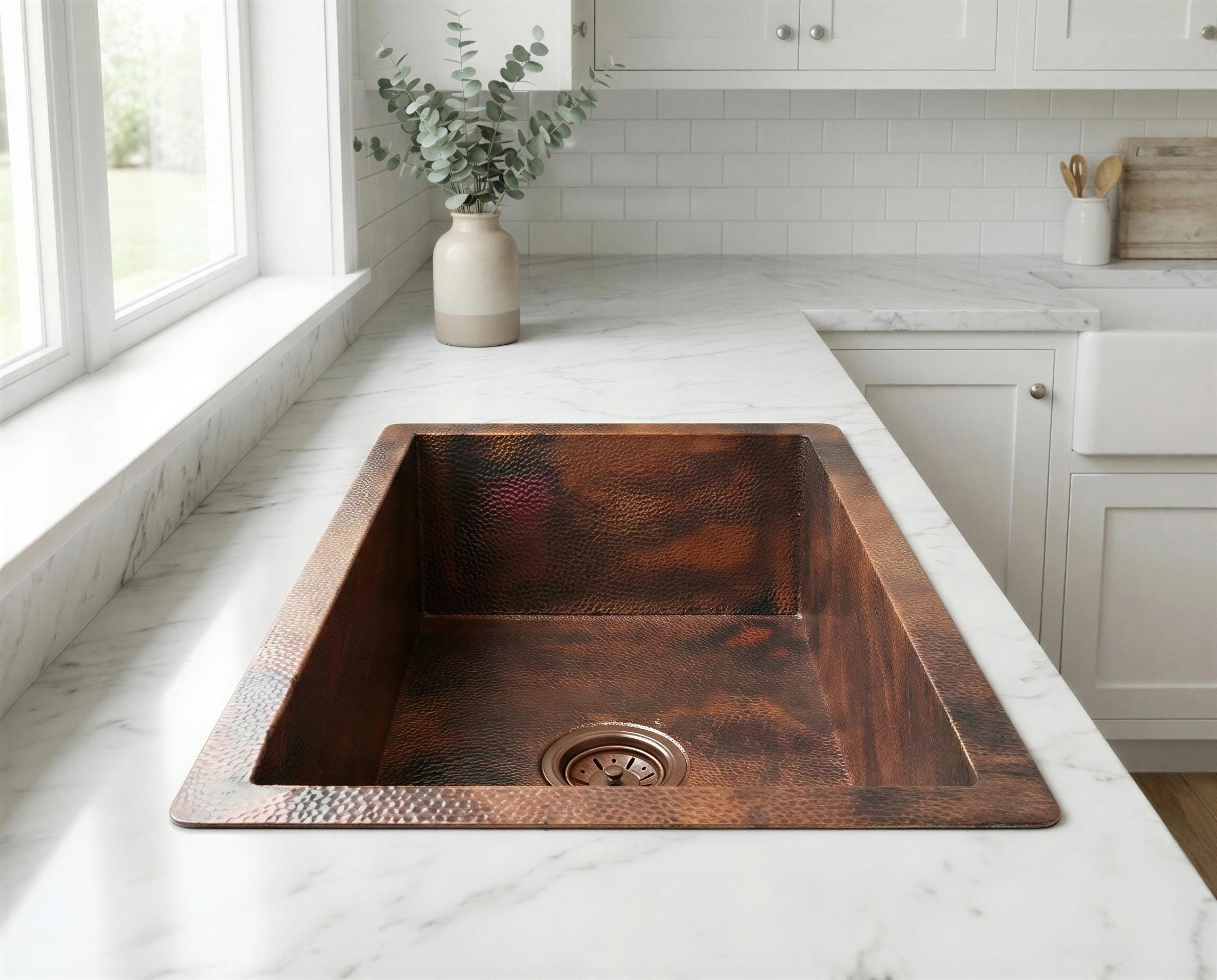 Copper kitchen sink on a marble countertop with a vase of greenery in the background.