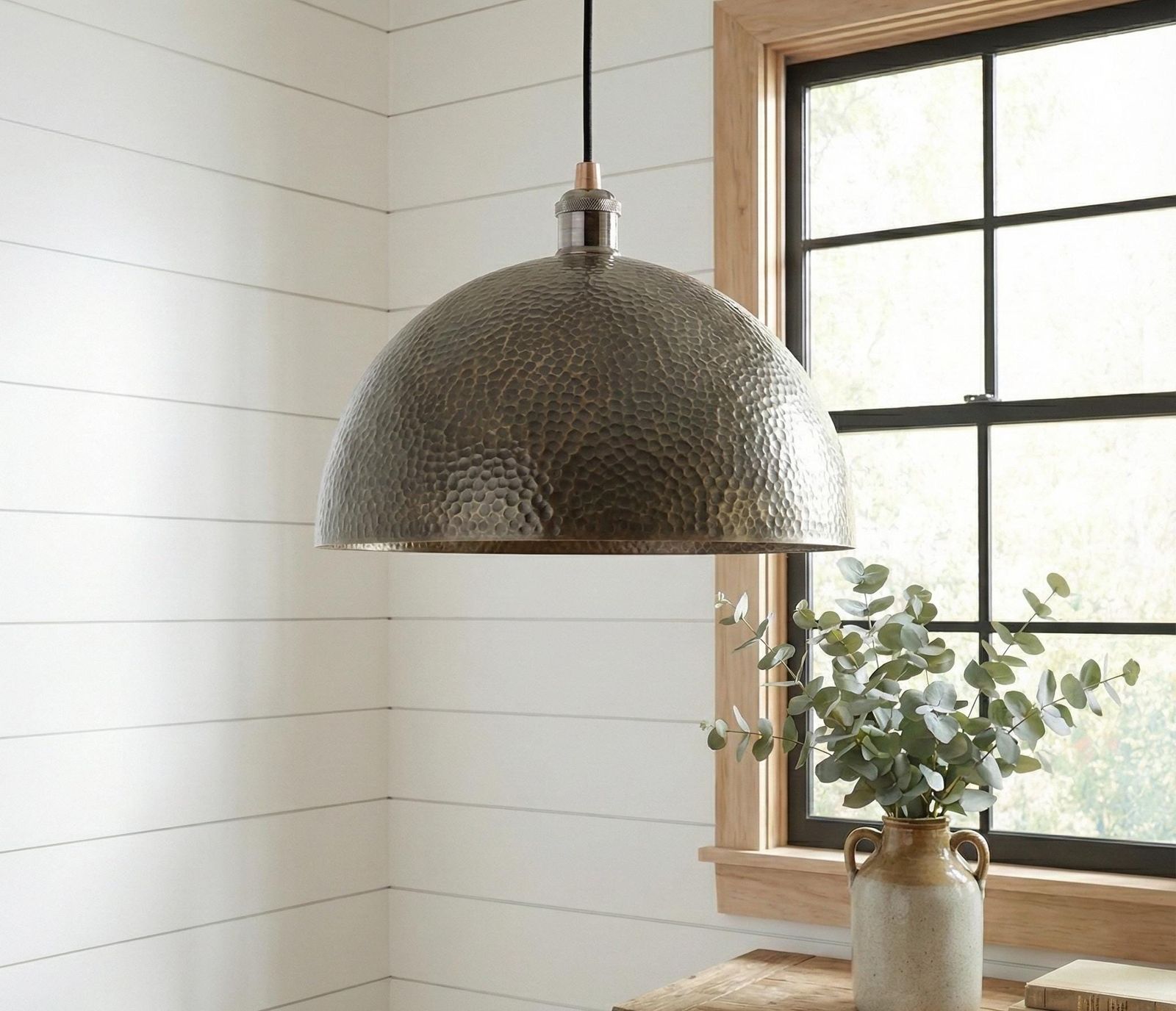 Hammered metal pendant light hanging in a room with a window and plant.