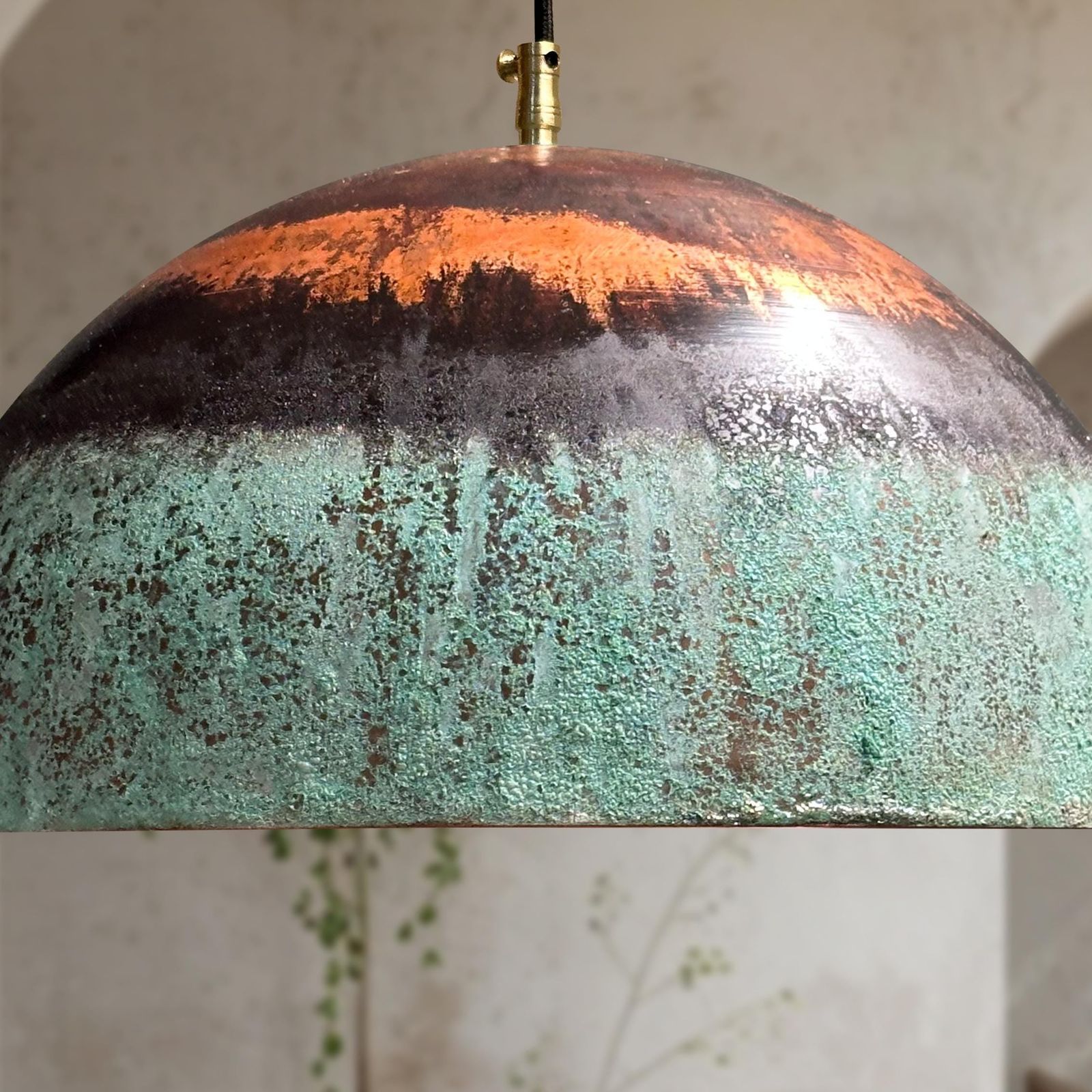 A close-up of a dome-shaped pendant light shade with a distressed, oxidized finish showing a gradient from copper-orange at the top to dark gray and mint green at the bottom, suspended by a brass fixture.