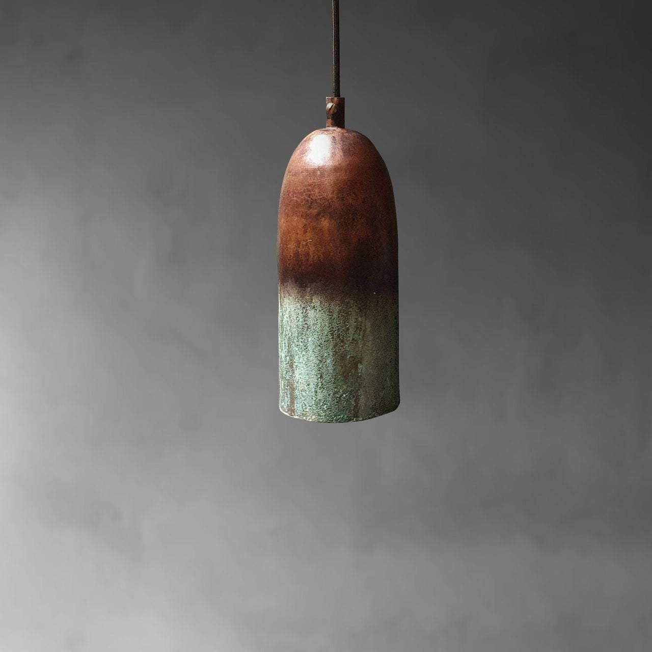 A single pendant light with a rounded, bullet-shaped shade featuring a rustic two-tone finish, transitioning from a warm copper-brown at the top to a textured greenish patina at the bottom, suspended against a dark gray background.