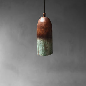 A single pendant light with a rounded, bullet-shaped shade featuring a rustic two-tone finish, transitioning from a warm copper-brown at the top to a textured greenish patina at the bottom, suspended against a dark gray background.