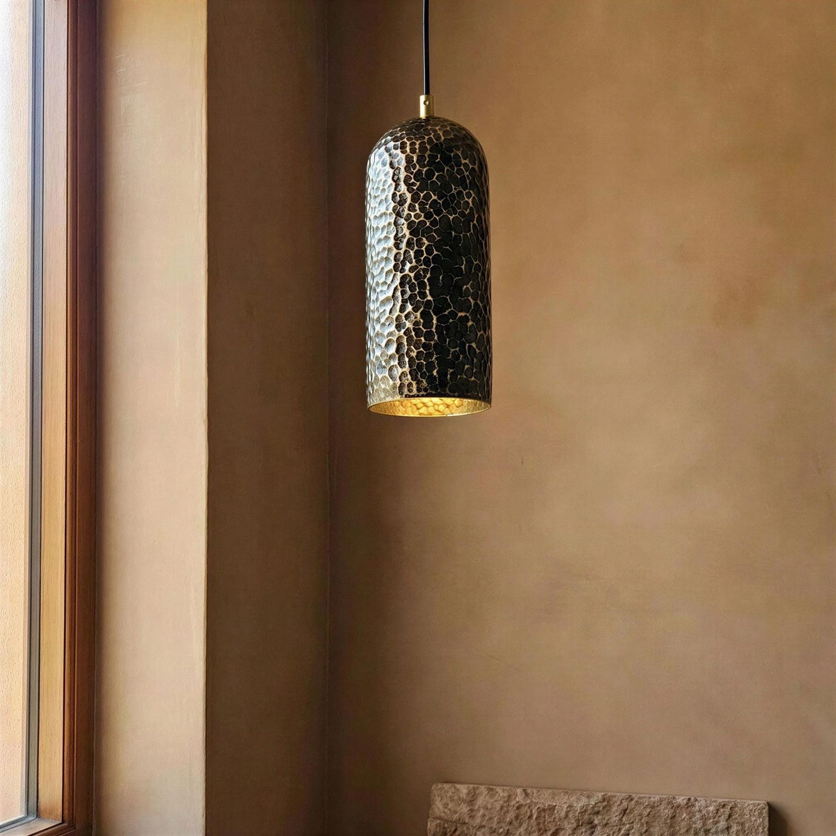 Hammered metal pendant light hanging against a brown wall with a window on the left.