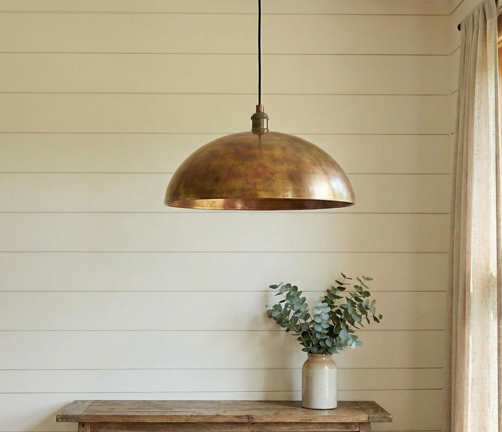 Bronze pendant light hanging above a wooden table with a vase of eucalyptus against a wooden paneled wall.