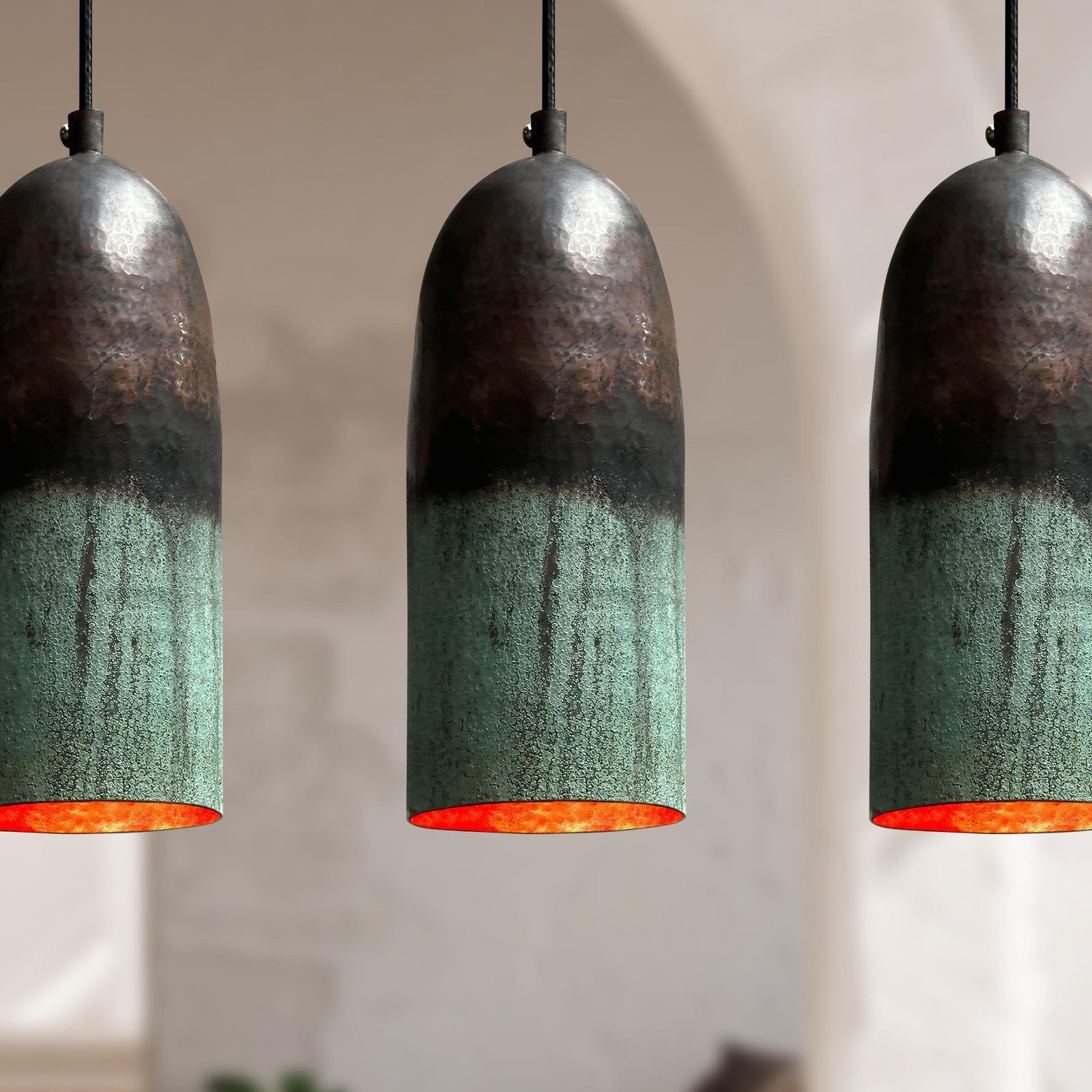 Three hanging pendant lights with metallic bronze upper sections and a textured green lower section, each showing a warm orange glow from the interior, set against a softly blurred beige background.