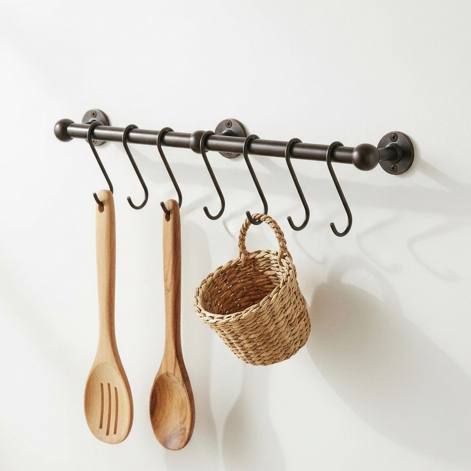 Wall-mounted metal rod with hooks holding wooden spoons and a woven basket on a white background
