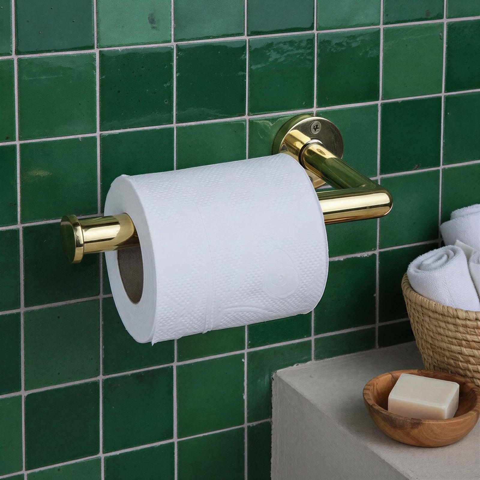 Gold toilet paper holder with a roll of toilet paper against a green tiled wall.