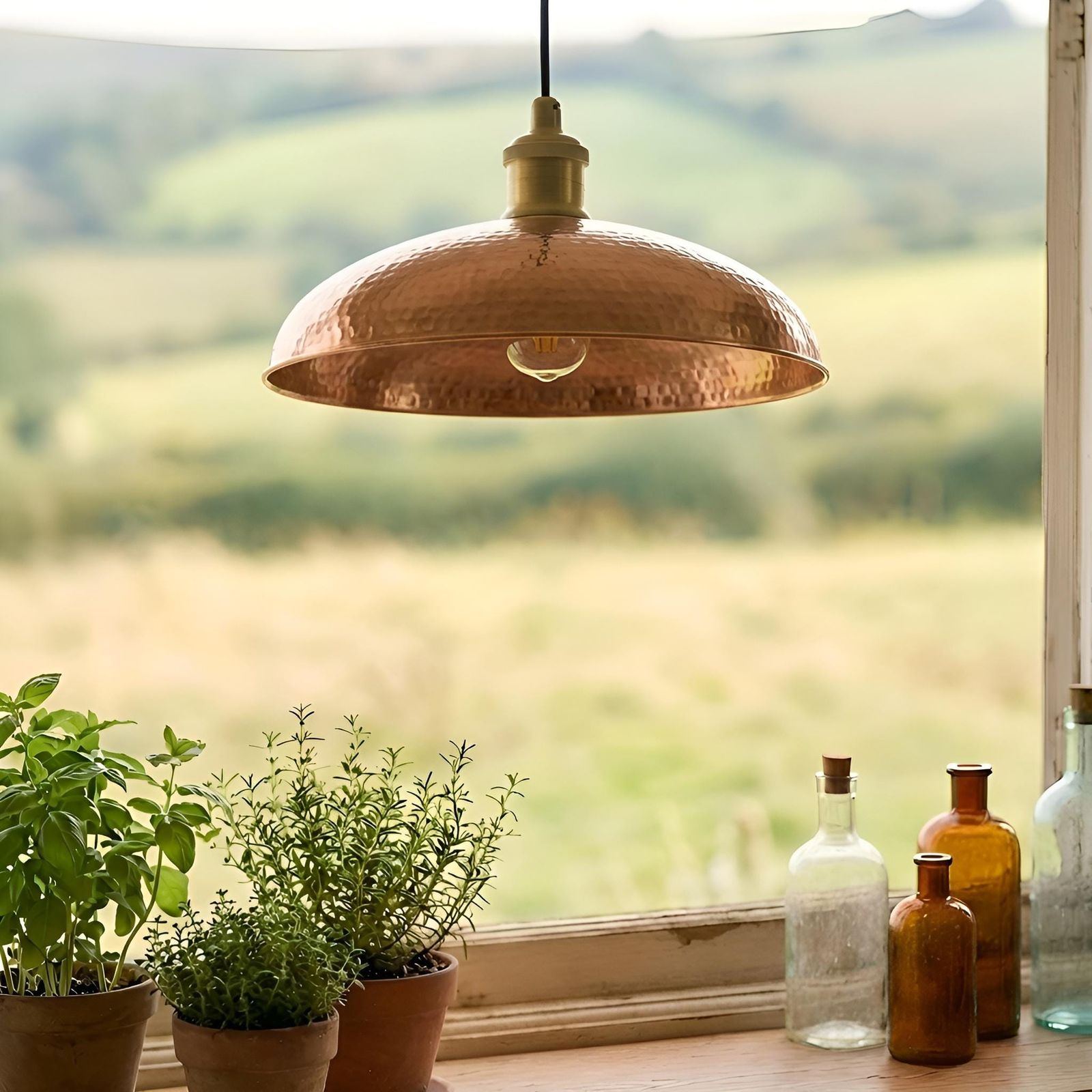 Copper pendant light hanging above a window with plants and bottles on a table.