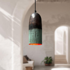 A single pendant light with a dark metallic, bullet-shaped shade featuring a green oxidized finish and a glowing orange-red interior, hanging in a softly blurred, sunlit modern interior with arches.
