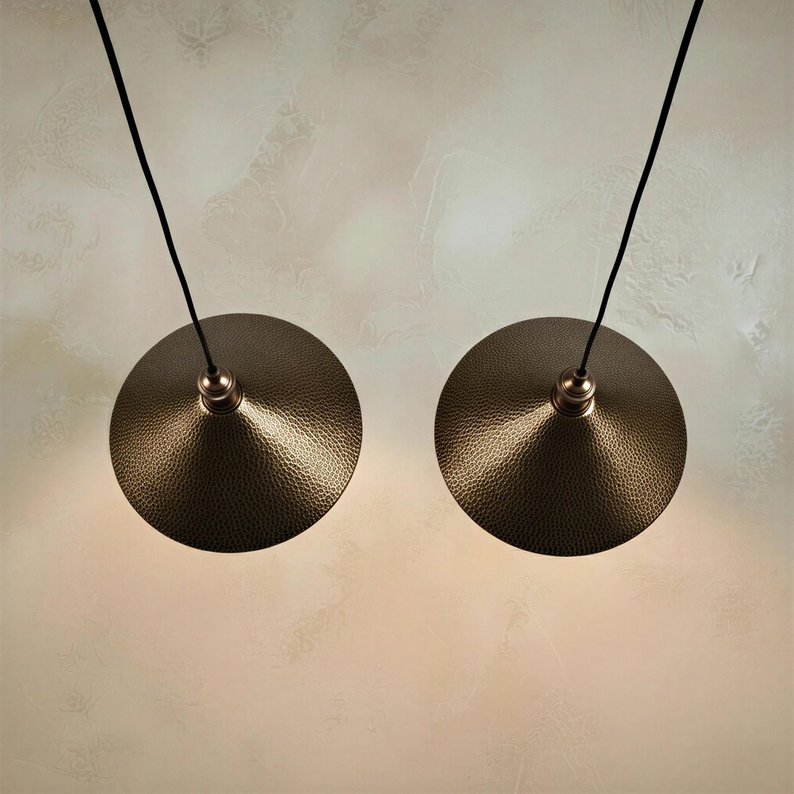 Rustic hammered brass hanging lamp in a flared funnel shape, casting soft illumination down against a warm beige background.