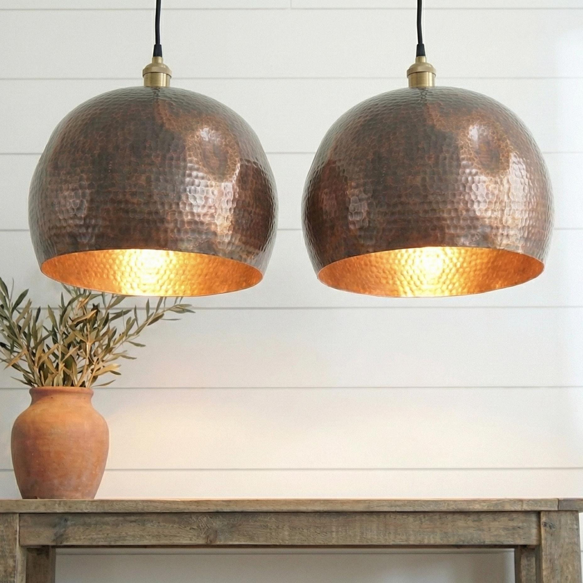 Two copper pendant lights hanging above a wooden table with a plant on a white paneled wall background.