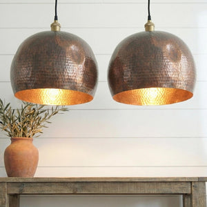 Two copper pendant lights hanging above a wooden table with a plant on a white paneled wall background.