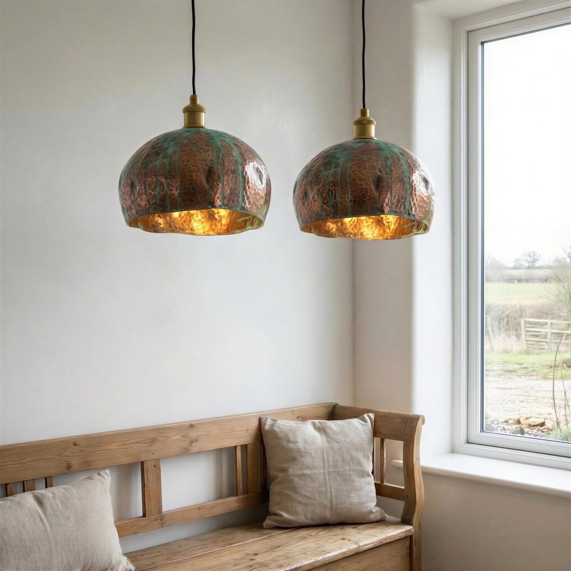 Two copper pendant lights hanging above a wooden bench with pillows in a room with a window.