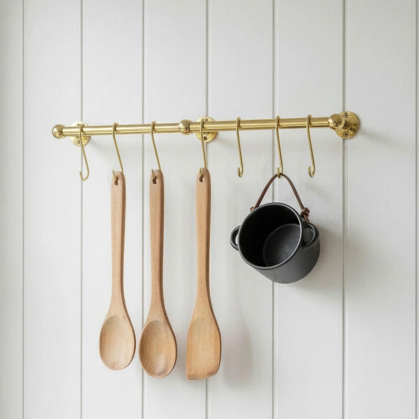Wooden spoons and a black pot hanging on a brass rod against a white paneled wall.