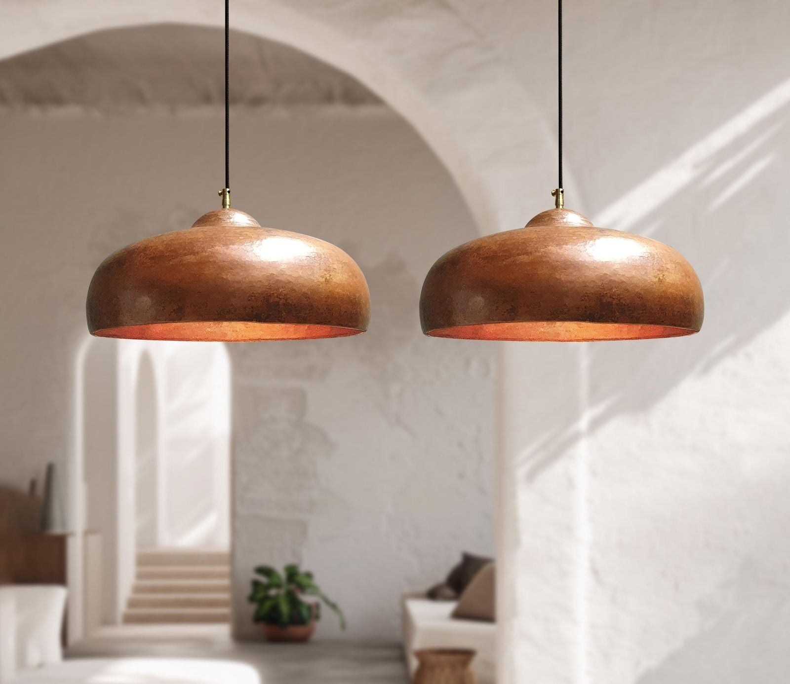 Two Copper pendant lights hanging in a room with a white wall and plant in the background.
