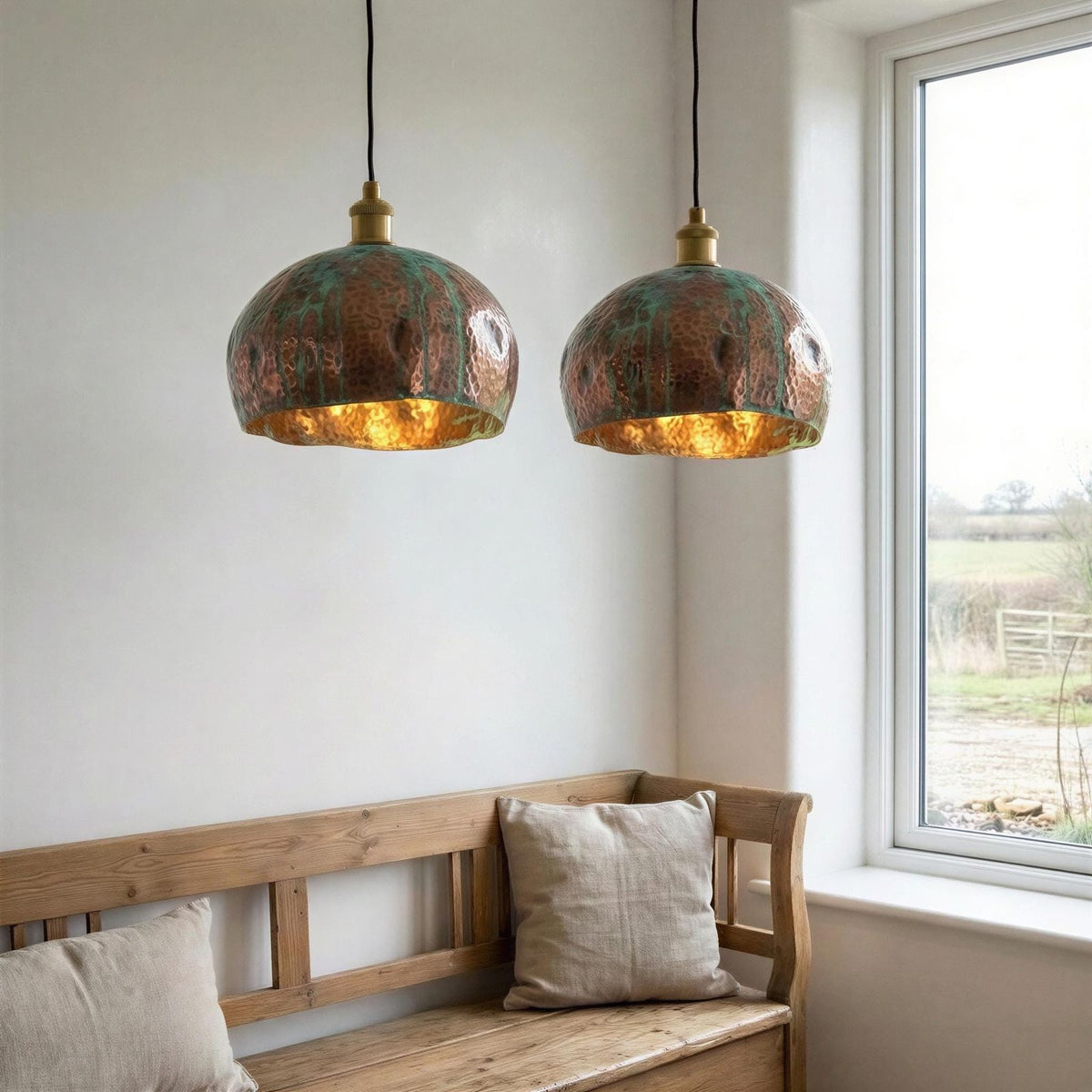 Two copper pendant lights hanging above a wooden bench with pillows in a room with a window.