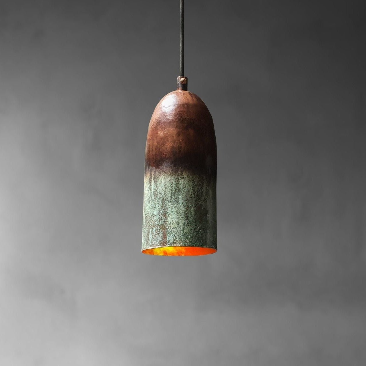 A single pendant light with a bullet-shaped shade made of oxidized metal, showing a dark brown top fading to a greenish patina at the bottom, with a glowing orange interior, suspended against a dark gray background.