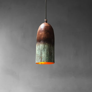 A single pendant light with a bullet-shaped shade made of oxidized metal, showing a dark brown top fading to a greenish patina at the bottom, with a glowing orange interior, suspended against a dark gray background.