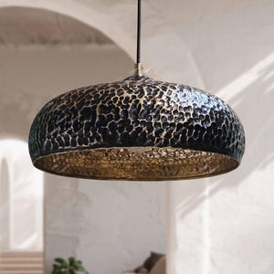 Wabi-sabi style dimpled brass hanging lamp positioned against blurred architectural archways and soft sunlight.