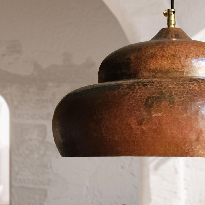 A close-up of a hammered copper pendant light shade with a brass ceiling mount, set against a softly blurred rustic white wall background.