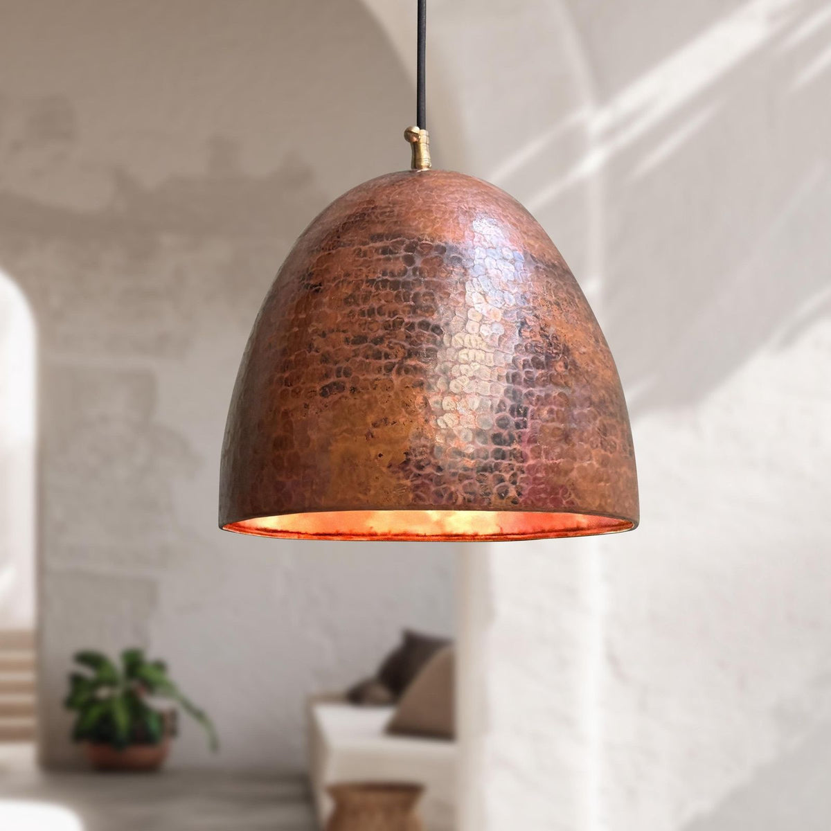 A hammered copper dome pendant light hanging in a softly lit interior with white walls and a blurred plant in the background.