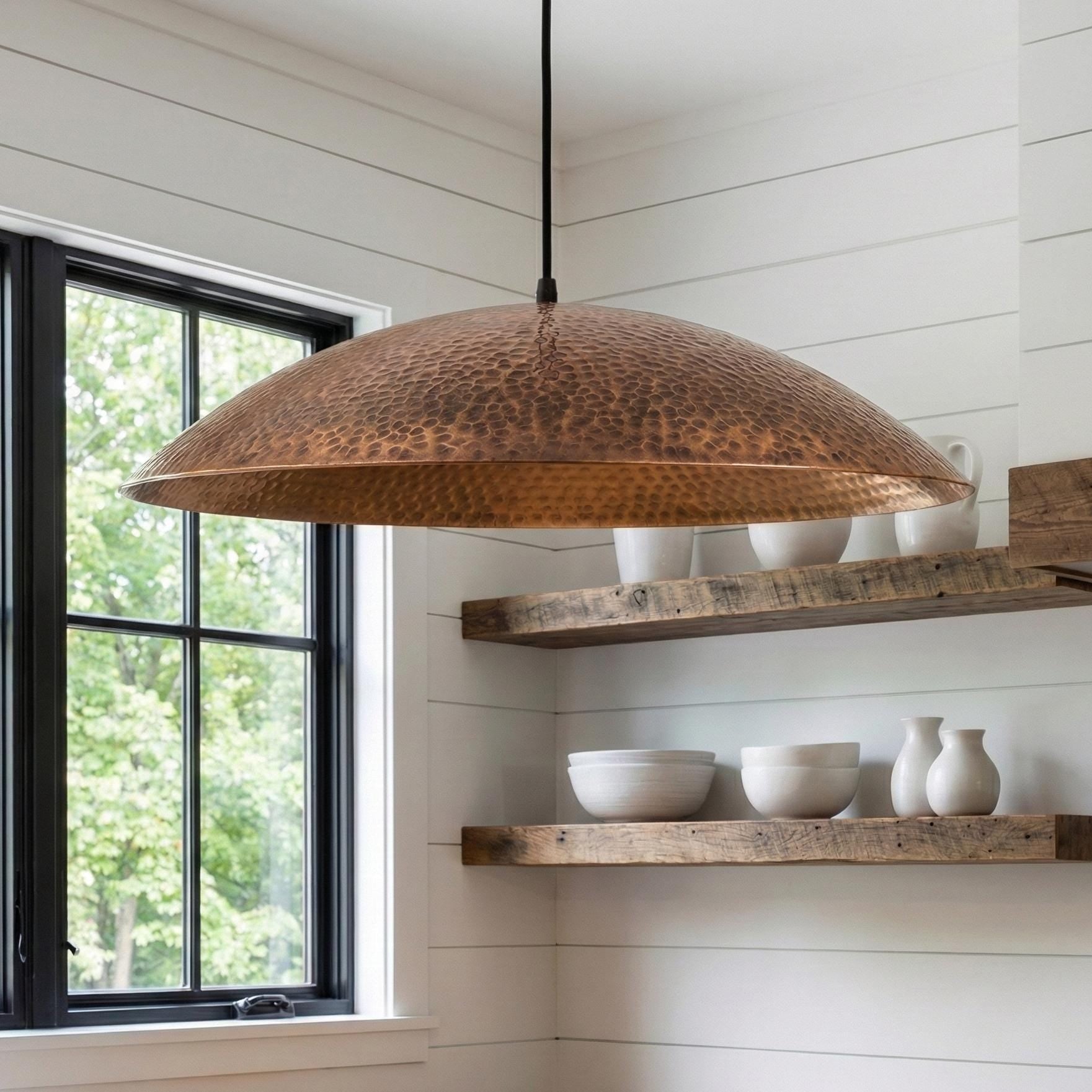 Copper pendant light hanging above wooden shelves with white bowls and vases in a room with large windows.