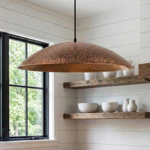 Copper pendant light hanging above wooden shelves with white bowls and vases in a room with large windows.