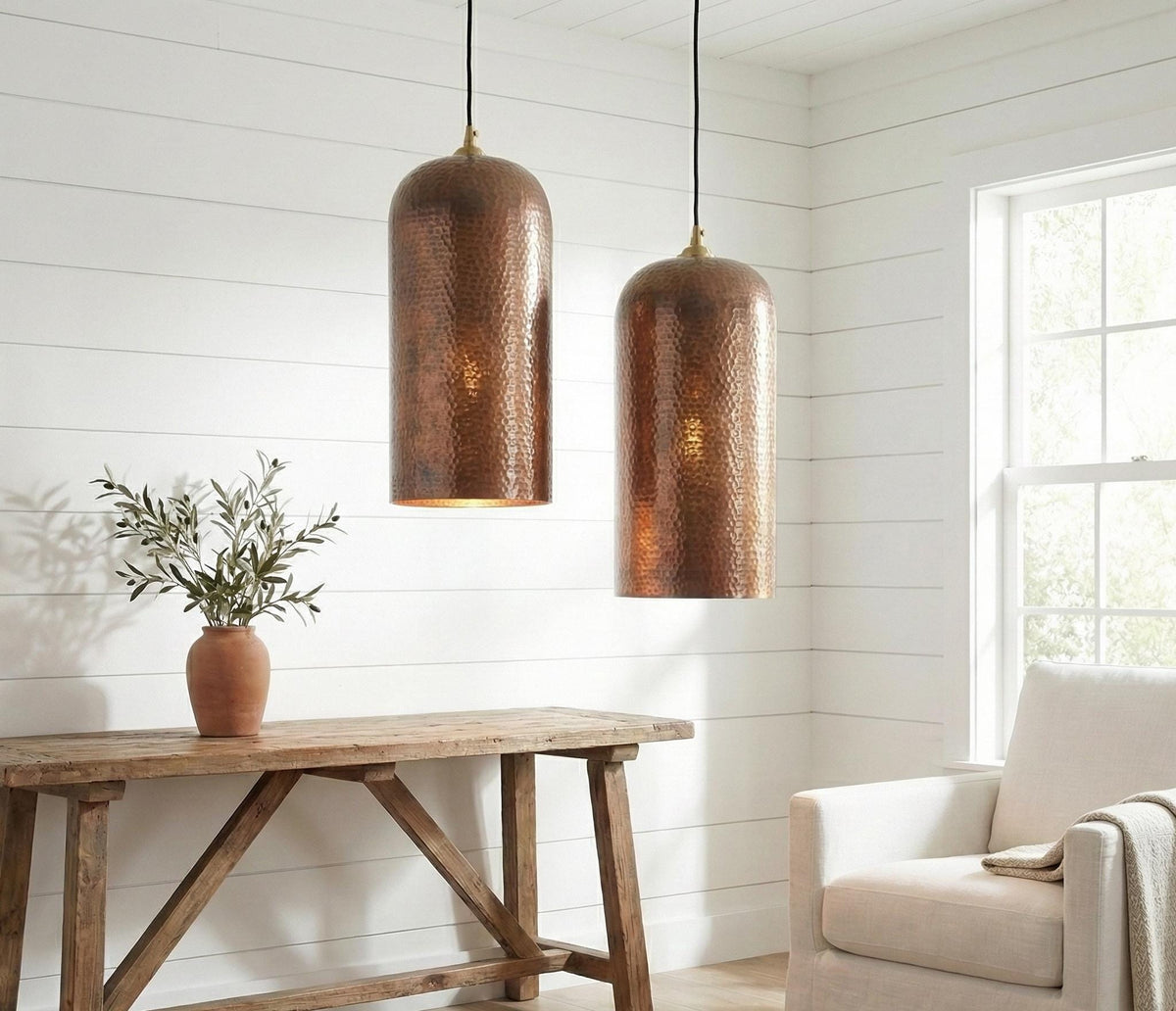Two copper pendant lights hanging above a wooden table with a plant and chair in a room with white walls.