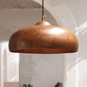 A hammered copper dome pendant light hanging from a black cord in a softly lit interior with white arches and plaster walls.
