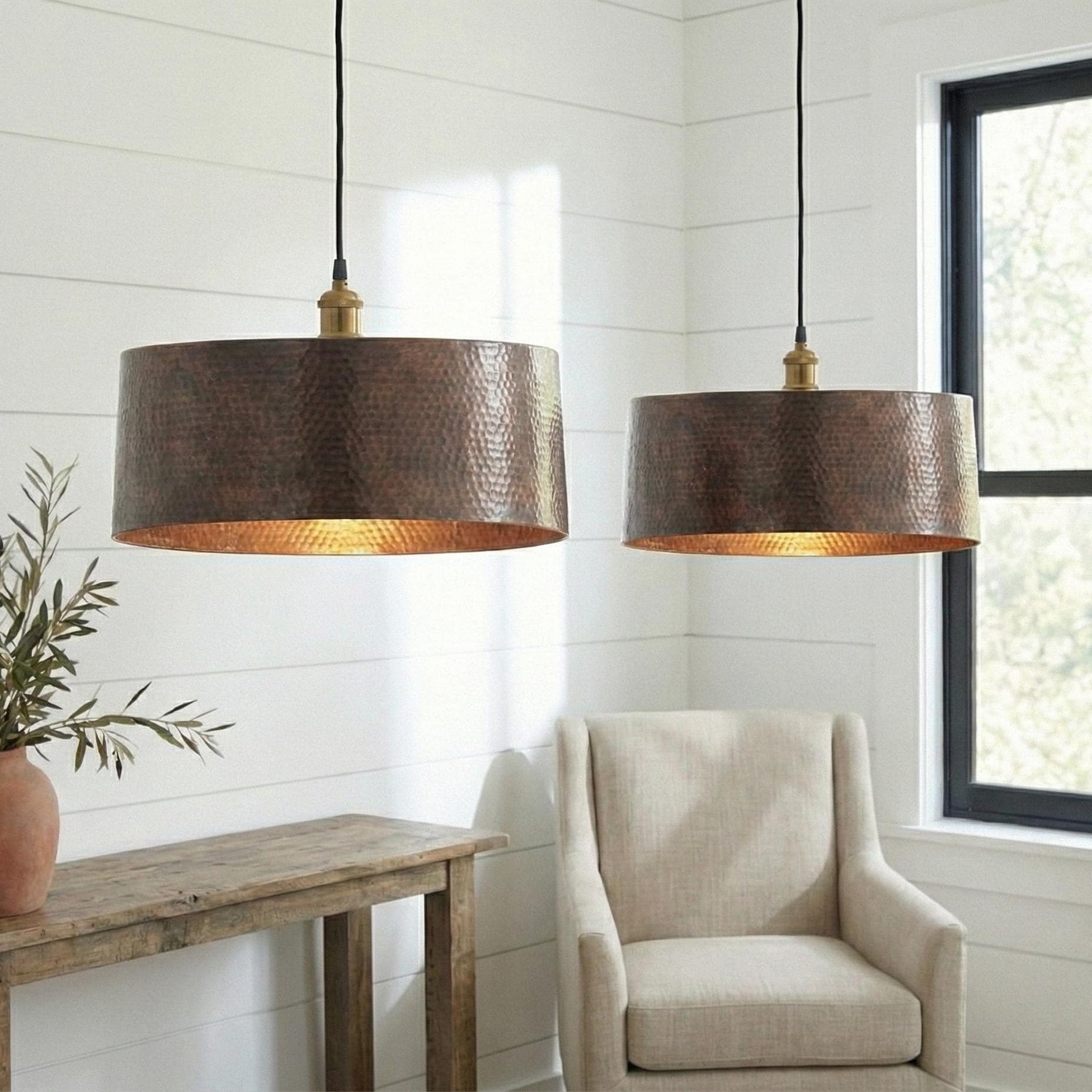 Two bronze pendant lights hanging above a chair and wooden table in a room with white walls and a window.