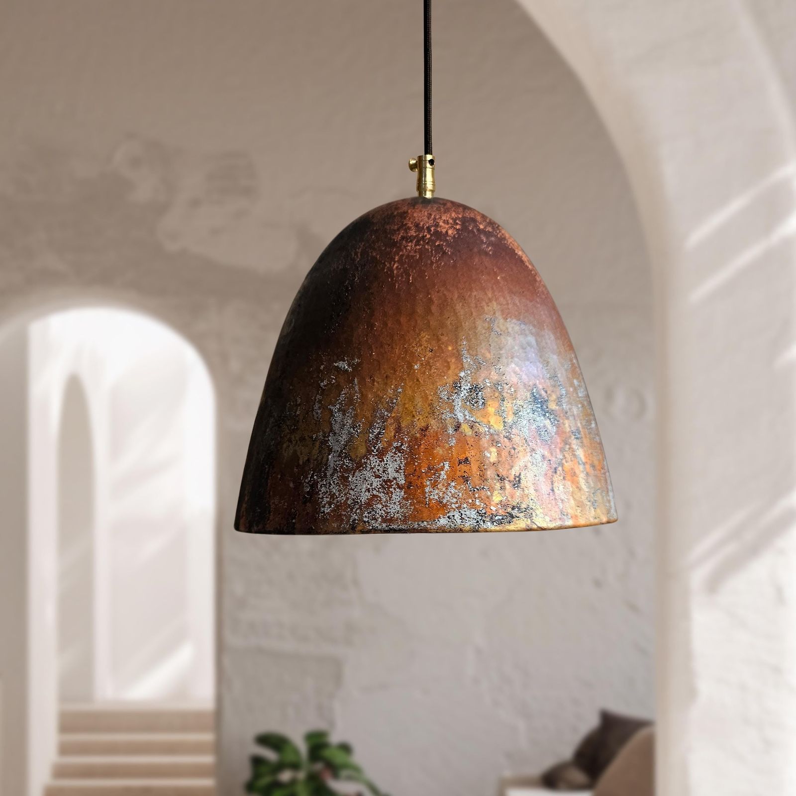 A dome-shaped pendant light with a distressed, hammered metallic finish in copper, bronze, and brown tones, hanging in a softly lit interior with arches and a blurred plant in the background.