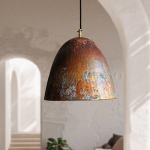 A dome-shaped pendant light with a distressed, hammered metallic finish in copper, bronze, and brown tones, hanging in a softly lit interior with arches and a blurred plant in the background.