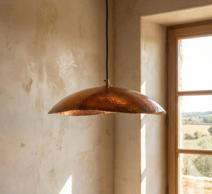 Copper pendant light hanging in a room with a window showing a view of trees.