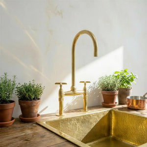 A polished brass bridge faucet with a high-arc spout and two handles, installed above a hammered brass sink on a rustic wooden countertop. The scene is styled with potted green herbs and a copper pot against a sunlit, textured off-white wall.