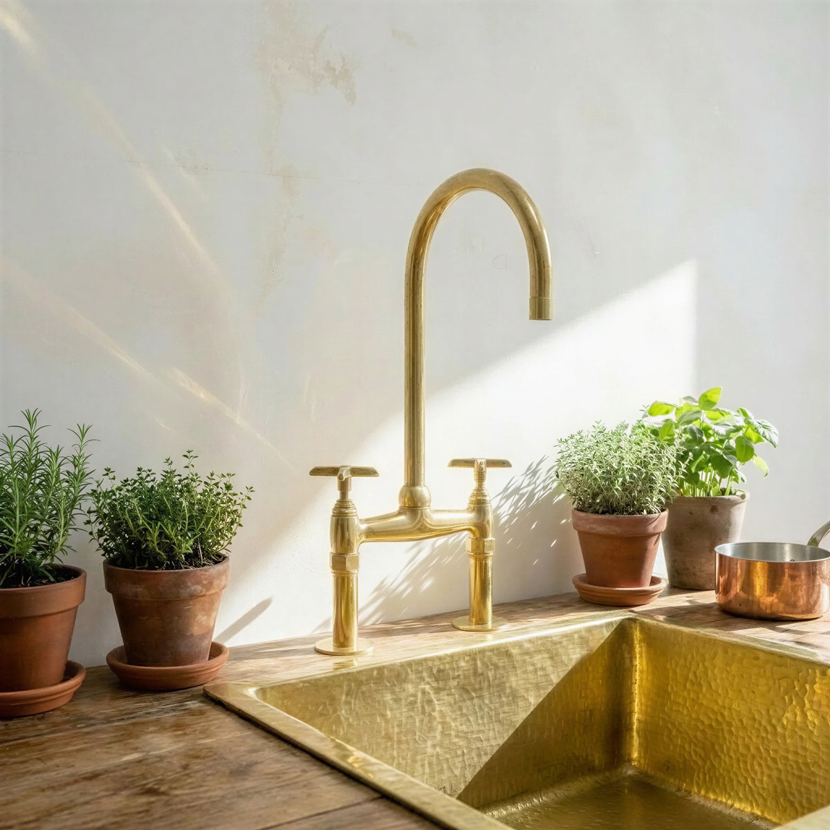A polished brass bridge faucet with a high-arc spout and two handles, installed above a hammered brass sink on a rustic wooden countertop. The scene is styled with potted green herbs and a copper pot against a sunlit, textured off-white wall.