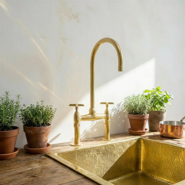 A polished brass bridge faucet with a high-arc spout and two handles, installed above a hammered brass sink on a rustic wooden countertop. The scene is styled with potted green herbs and a copper pot against a sunlit, textured off-white wall.