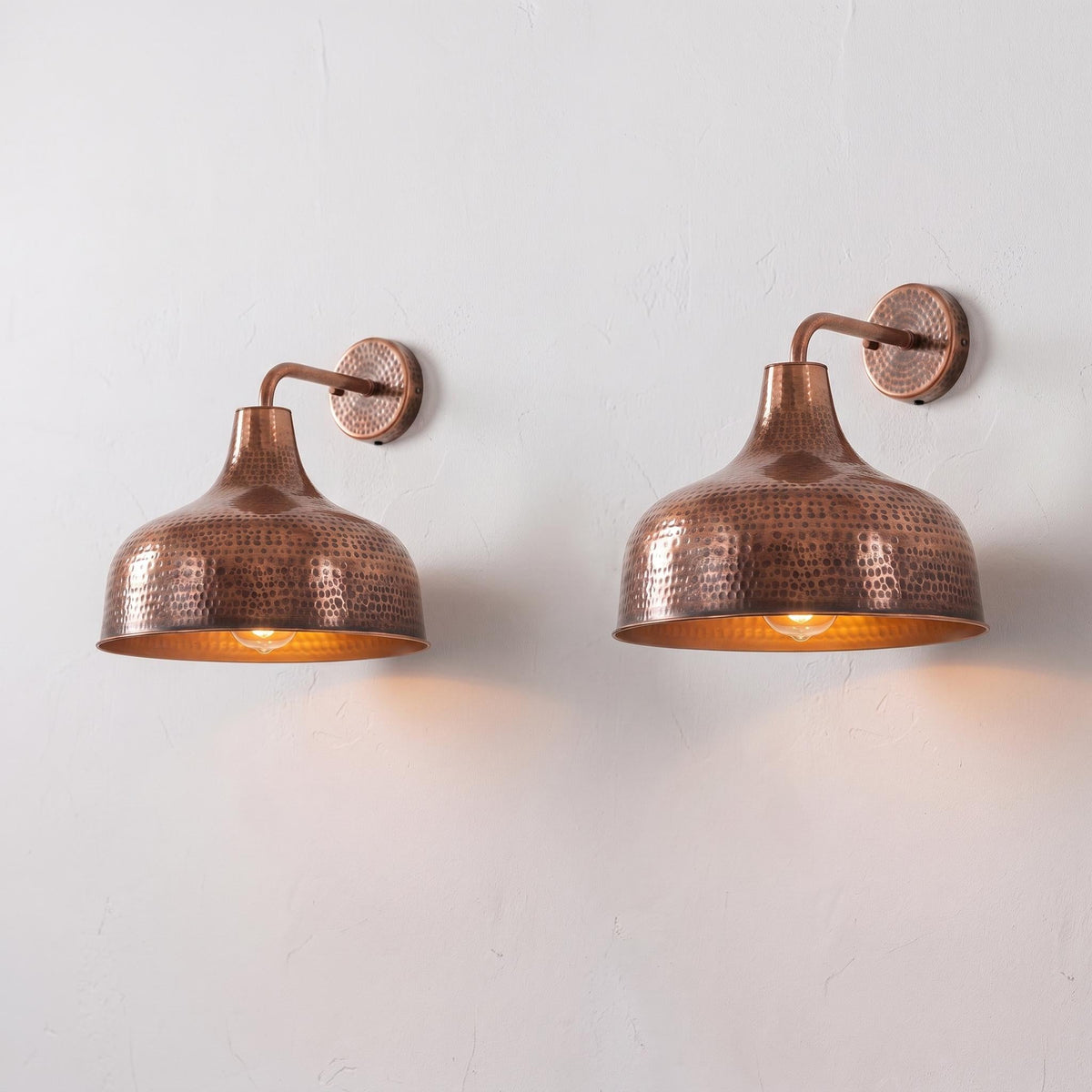Two matching hammered copper wall sconces with warm glowing bulbs, mounted on a light gray textured wall.