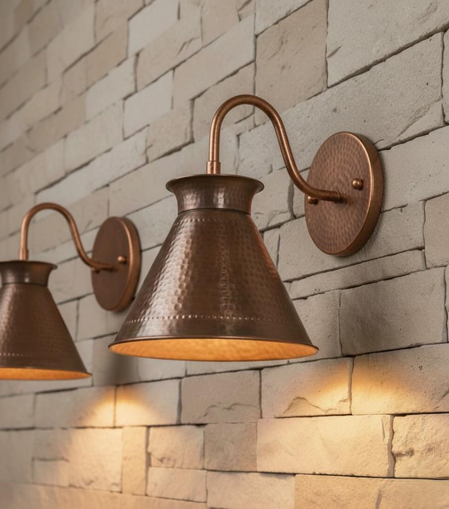Two copper wall sconces with hammered metal shades and curved arms mounted on a light beige stone brick wall, casting a warm yellow light.