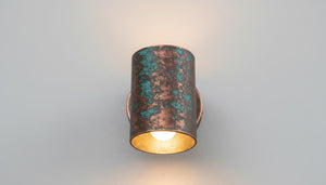 A cylindrical wall sconce with a distressed copper and turquoise patina finish, casting a warm light against a neutral gray background.
