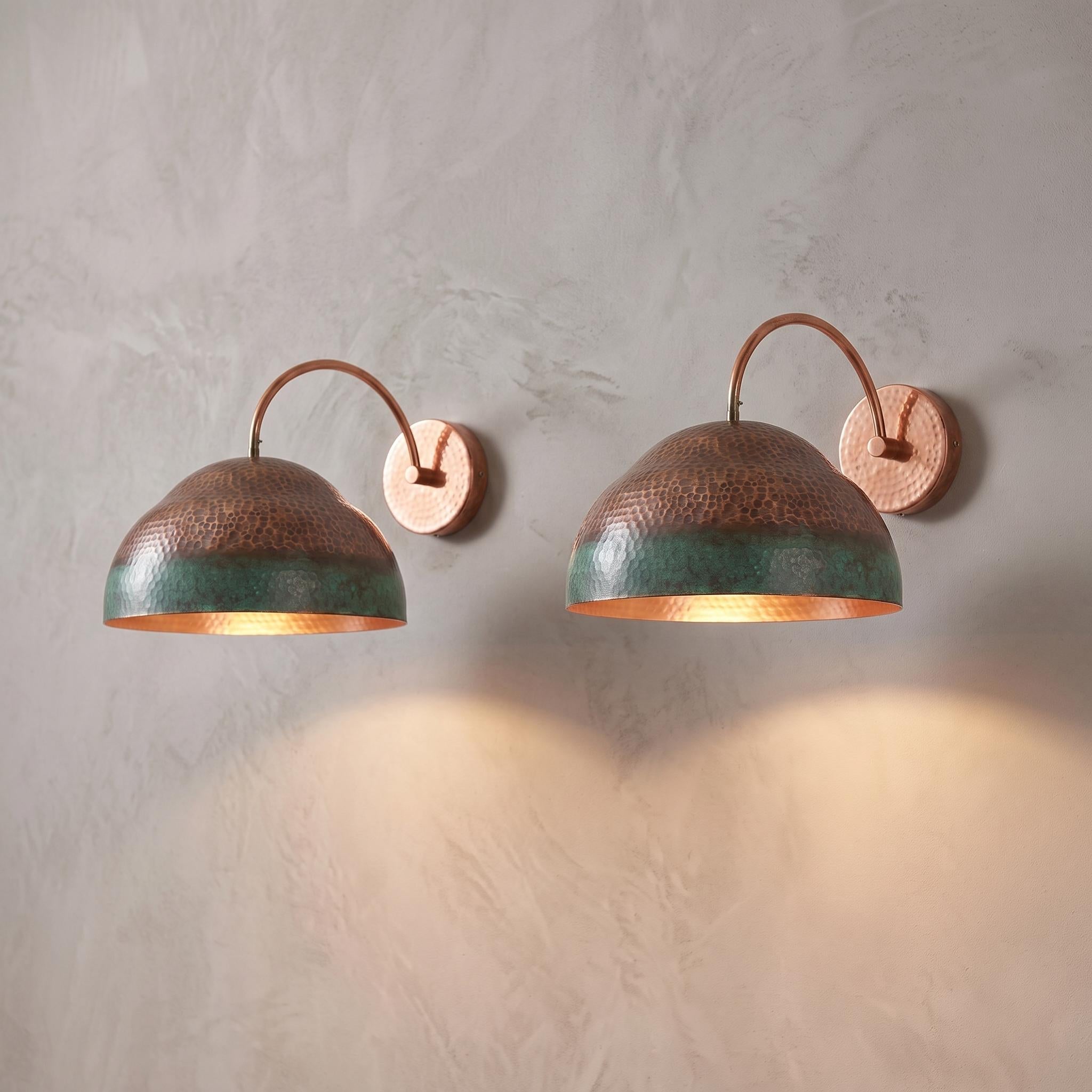 Two copper wall sconces with hammered dome shades and a green-blue patina lower band, mounted on a textured light gray plaster wall and casting warm light downward.