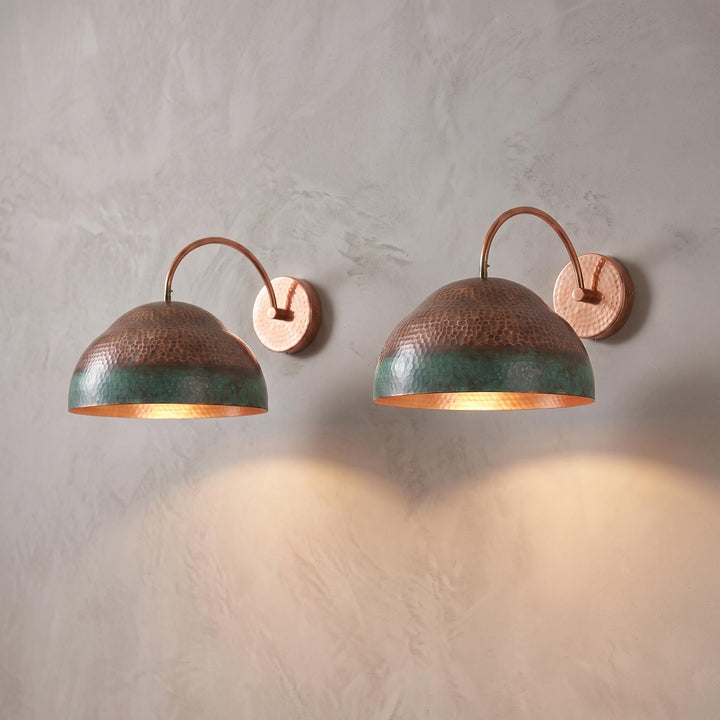 Two copper wall sconces with hammered dome shades and a green-blue patina lower band, mounted on a textured light gray plaster wall and casting warm light downward.